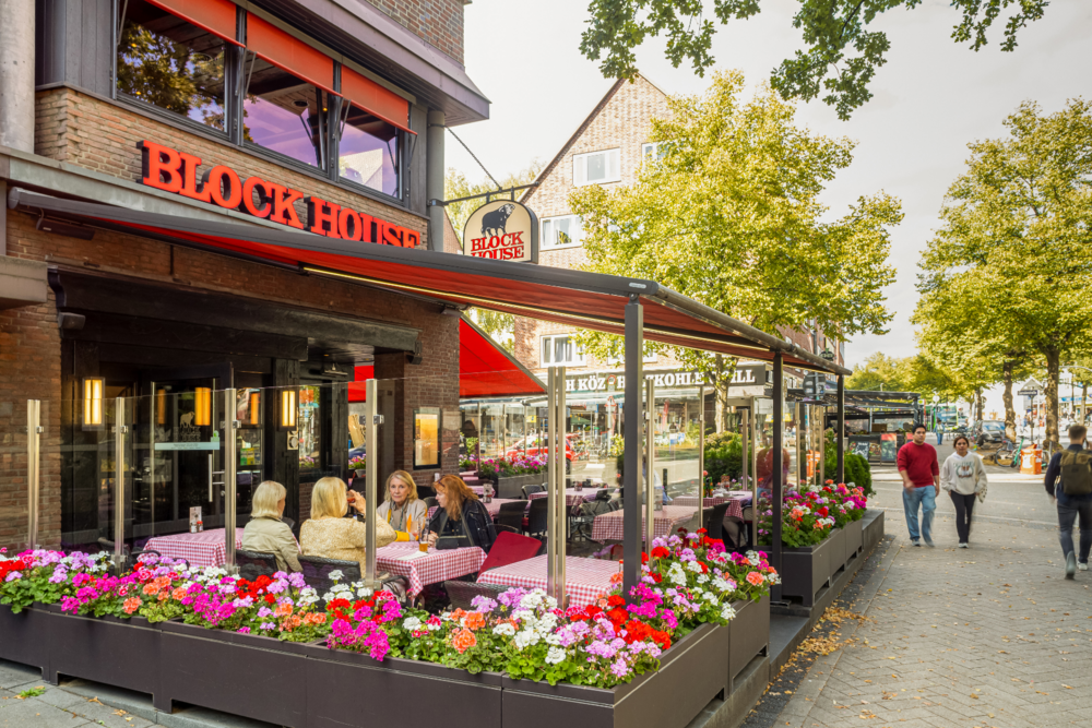 BLOCK HOUSE Hamburg Barmbek – sonnengeschützte Terrasse Terrasse des BLOCK HOUSE Hamburg Barmbek mit Gästen, rot-weiß karierten Tischdecken und Blumen vor dem Steakhouse