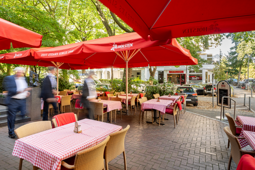 BLOCK HOUSE Winterhude Hamburg – Steakhouse mit Terrasse Außenansicht der Terrasse vom BLOCK HOUSE Hamburg-Winterhude mit gedeckten Tischen, roten Schirmen und großen Bäumen