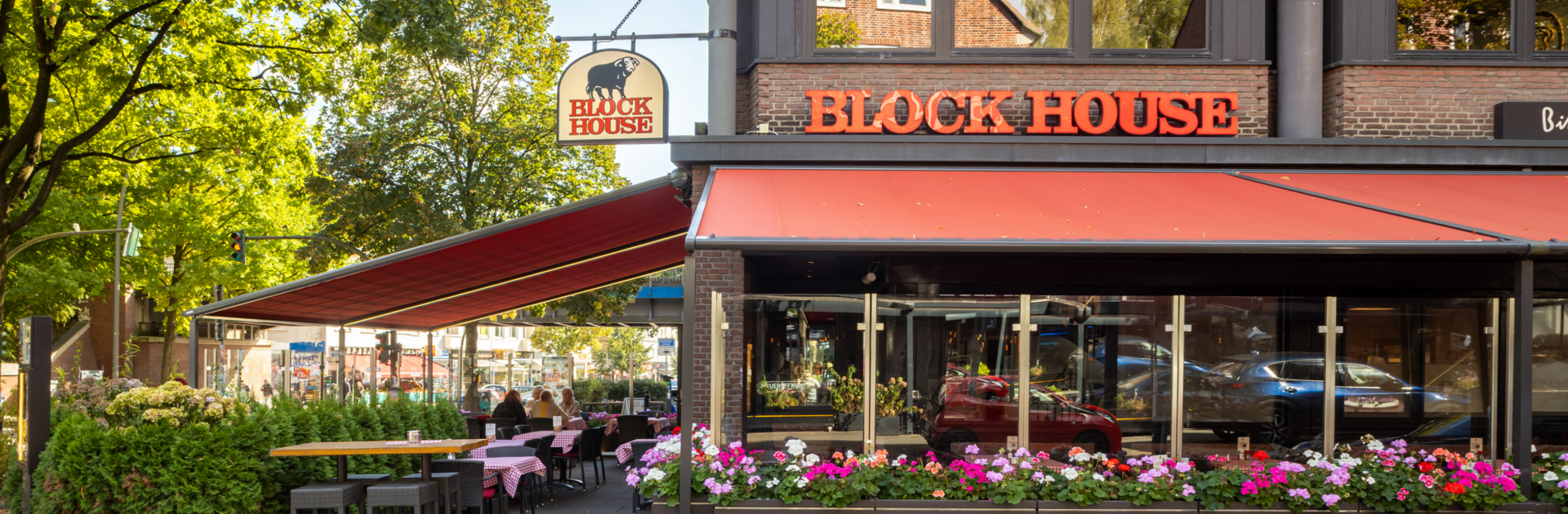 BLOCK HOUSE Hamburg Barmbek – Steakhouse mit Terrasse Außenansicht des BLOCK HOUSE Hamburg Barmbek mit Terrasse, roten Markisen und Sitzplätzen vor dem Steakhouse