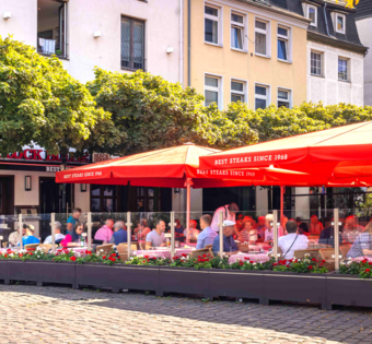 BLOCK HOUSE Restaurant Düsseldorf Burgplatz Außenansicht des BLOCK HOUSE Düsseldorf am Burgplatz mit großer Terrasse, roten Sonnenschirmen und Gästen.