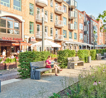 Blick auf das BLOCK HOUSE Restaurant in Ahrensburg. Im Vordergrund sind ein gepflasterter Weg mit grünen Bäumen und Sitzbänken zu sehen. 