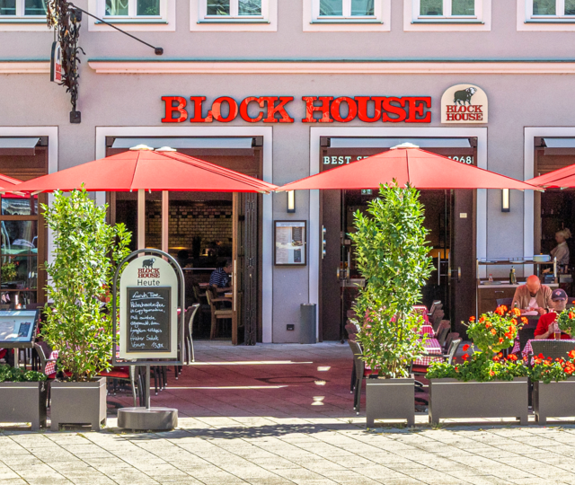 BLOCK HOUSE Restaurant Augsburg Außenansicht des BLOCK HOUSE Restaurants in Augsburg: eine große, hölzerne Fensterfront, davor viele Sitzgelegenheiten auf der Terrasse, die sich direkt auf dem Ulrichsplatz befindet.