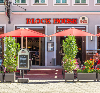 BLOCK HOUSE Restaurant Augsburg Außenansicht des BLOCK HOUSE Restaurants in Augsburg: eine große, hölzerne Fensterfront, davor viele Sitzgelegenheiten auf der Terrasse, die sich direkt auf dem Ulrichsplatz befindet.