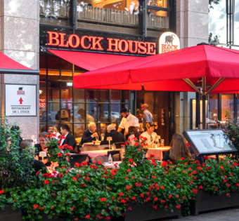 BLOCK HOUSE München Elisenhof Außenansicht des BLOCK HOUSE München Elisenhof mit Terrasse, roten Sonnenschirmen und Gästen im Außenbereich.