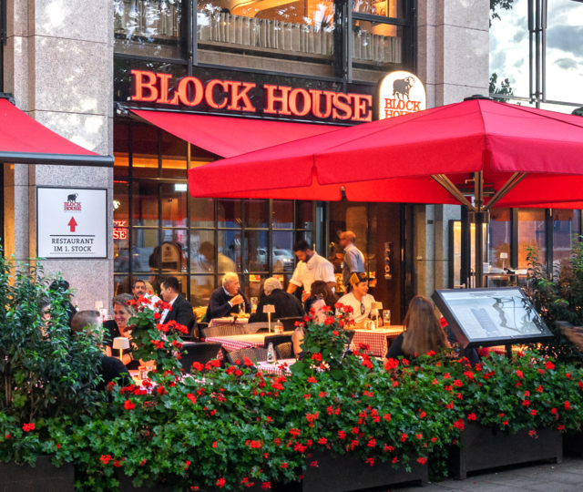 BLOCK HOUSE München Elisenhof Außenansicht des BLOCK HOUSE München Elisenhof mit Terrasse, roten Sonnenschirmen und Gästen im Außenbereich.