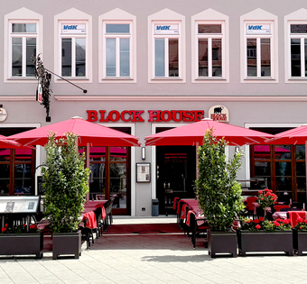 BLOCK HOUSE Restaurant Augsburg Außenansicht des BLOCK HOUSE Restaurants in Augsburg: eine große, hölzerne Fensterfront, davor viele Sitzgelegenheiten auf der Terrasse, die sich direkt auf dem Ulrichsplatz befindet.