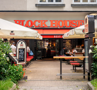 BLOCK HOUSE München Leopoldstraße – Steakhouse mit Terrasse Eingang und Terrasse des BLOCK HOUSE München Leopoldstraße mit Sonnenschirmen und Gästen vor dem Steakhouse