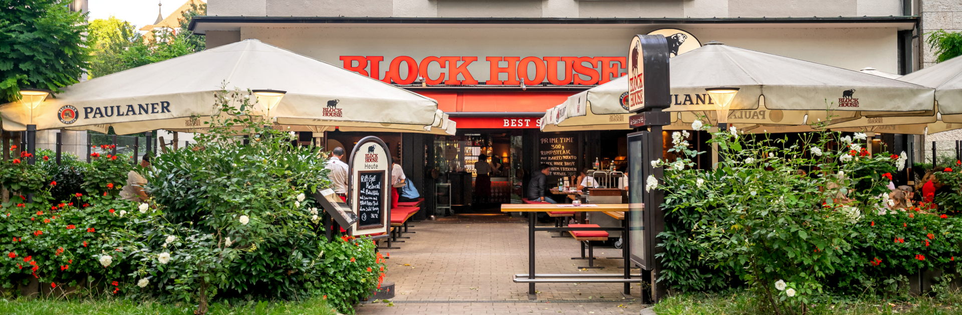 BLOCK HOUSE München Leopoldstraße – Steakhouse mit Terrasse Eingang und Terrasse des BLOCK HOUSE München Leopoldstraße mit Sonnenschirmen und Gästen vor dem Steakhouse